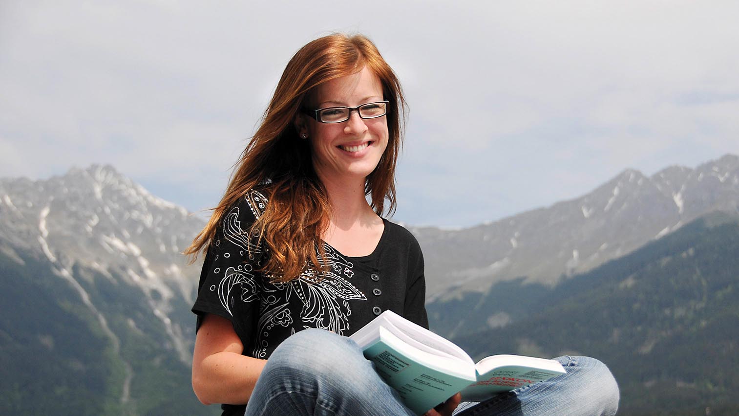 Young female student sitting cross-legged with book, a mountain range in the background